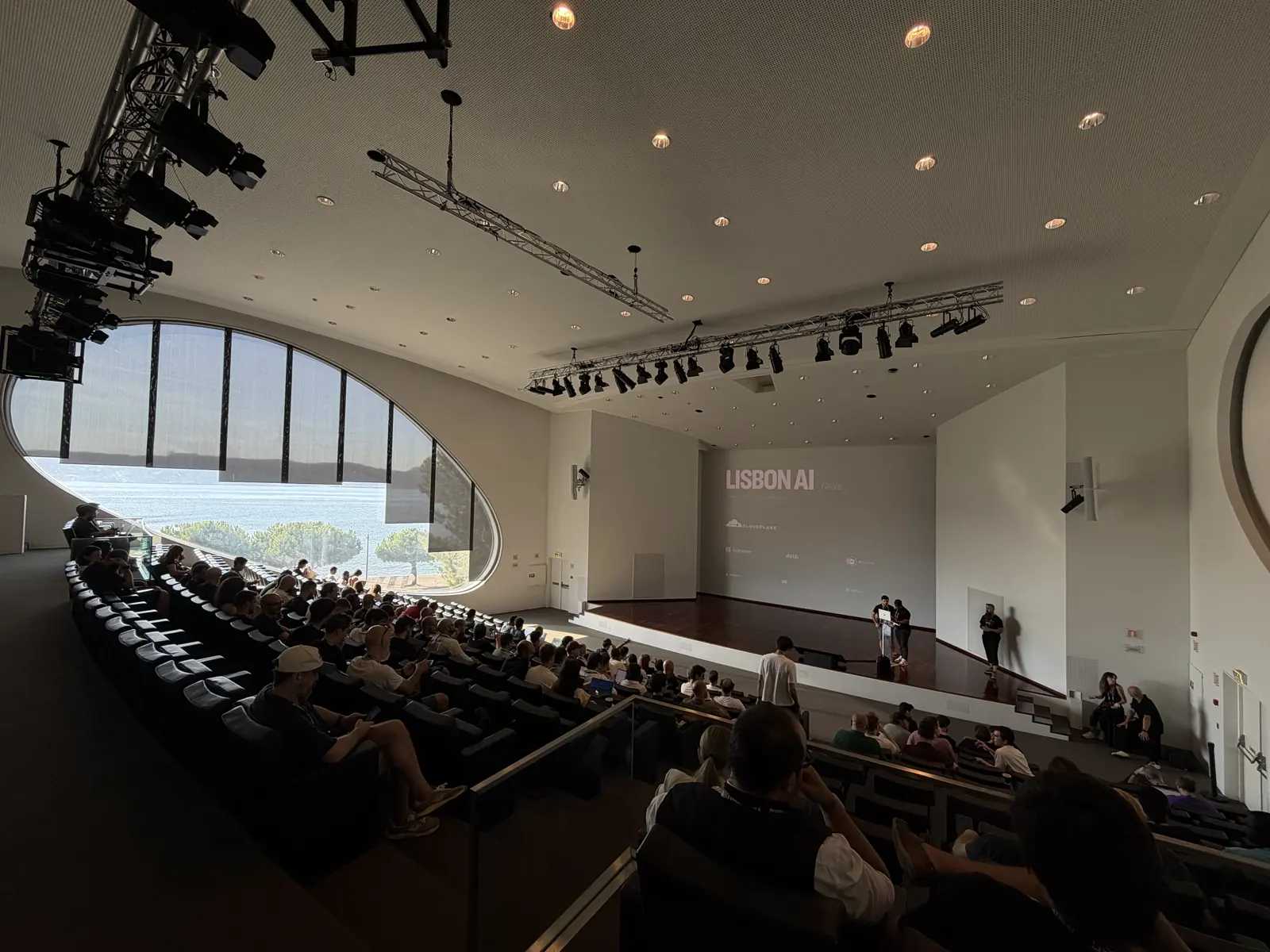 Nearly full tiered auditorium, with a large oval window on the left and the stage at the back.