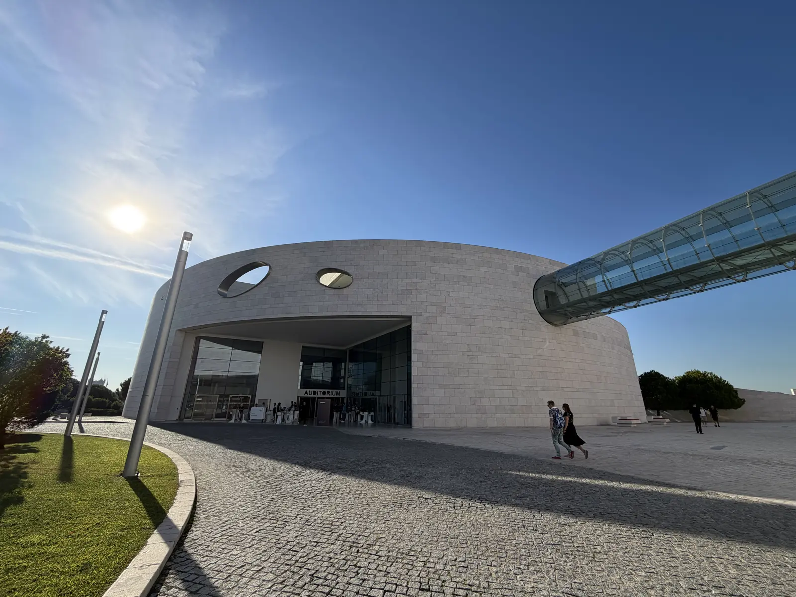 Curved facade of the Champalimaud Centre under a blue sky, with a glass walkway coming out of the building and the sun overhead.