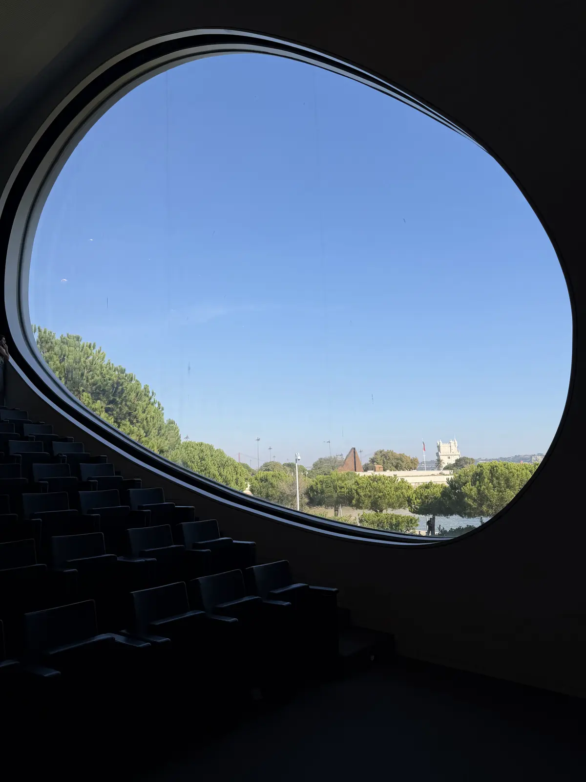 The oval window seen from inside the dark auditorium, framing trees and blue sky.