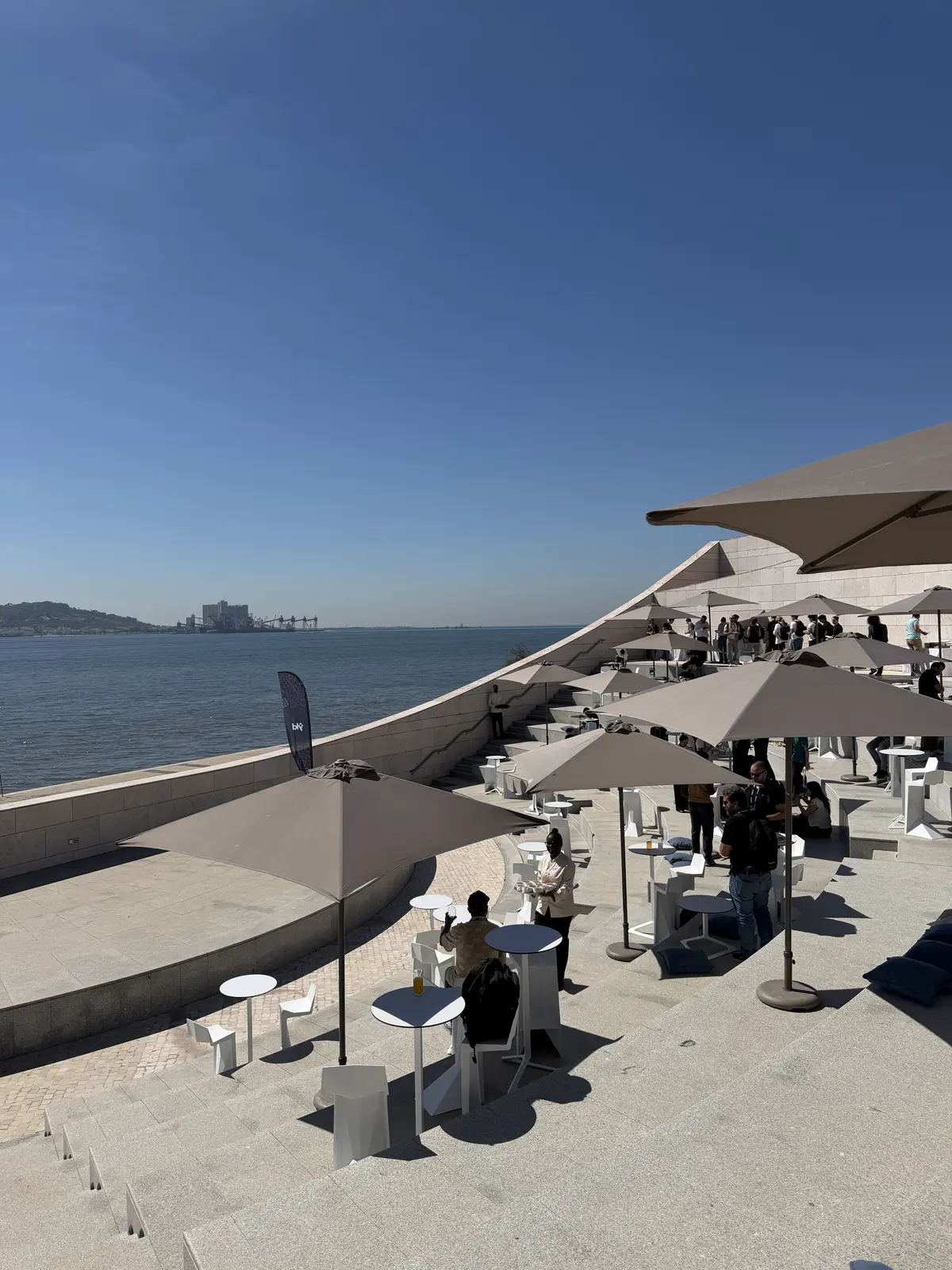 Terrace with umbrellas and tables full of people, with the river in the background.